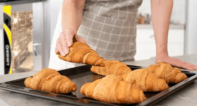 cruasanes tostados con una técnica de cocción en hornos de convección y siendo colocados en una charola por un panadero en primer plano con un horno de conveccion al fondo