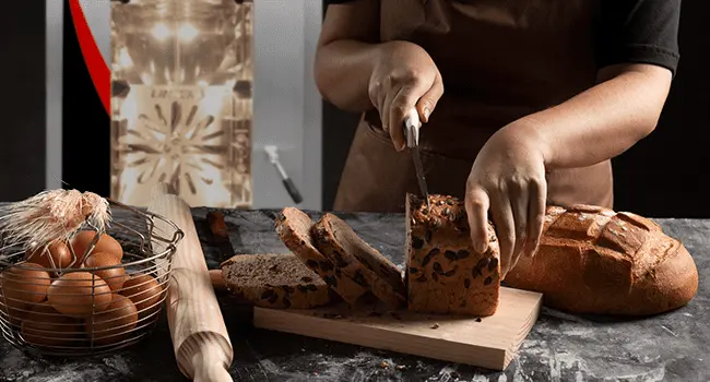 pan horneado con una técnica de cocción en hornos de convección y siendo cortado por un panadero en primer plano con un horno de conveccion al fondo