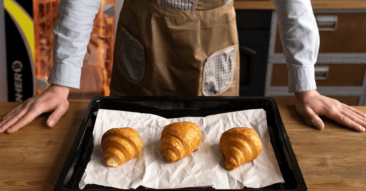 Panadero mostrando sus croissants sobre una bandeja para hornear con un horno LINHER de fondo