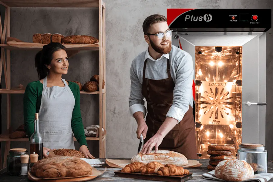 Pareja de panaderos cortando pan en una cocina de panadería con un horno de convección al fondo