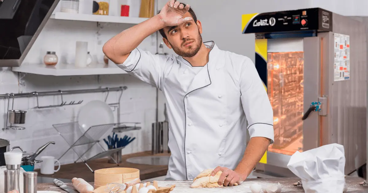 Hombre panadero agotado en una cocina de panadería con un horno de convección Goliat de fondo