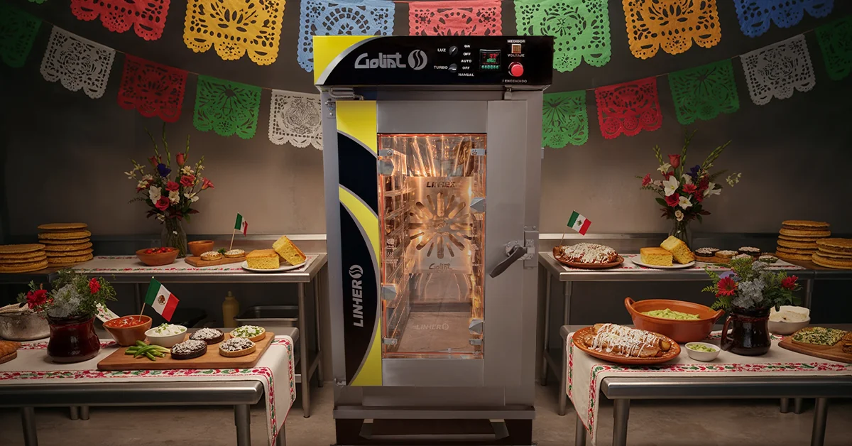 Cocina profesional mexicana decorada con papel picado tricolor y banderas, con un horno de convección LINHER Goliat en primer plano y charolas de sopes, enchiladas con mole y pan de elote. El ambiente transmite orgullo patrio, celebración y profesionalismo.