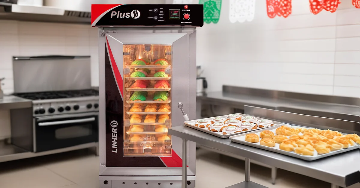 Horno de convección abierto en primer plano con conchas tricolor, roles glaseados con franjas verde y roja y panes de atole; papel picado tricolor al fondo y mesa de acero con charolas listas, luz cálida.